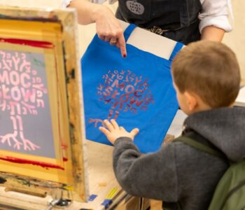 chłopiec dotyka torby wykonanej metodą sitodruku. Nadruk przedstawia drzewko z napisem: Moc sł&oacute;w