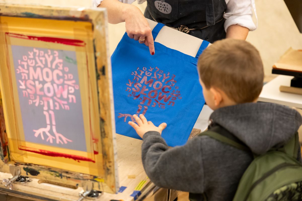 chłopiec dotyka torby wykonanej metodą sitodruku. Nadruk przedstawia drzewko z napisem: Moc słów